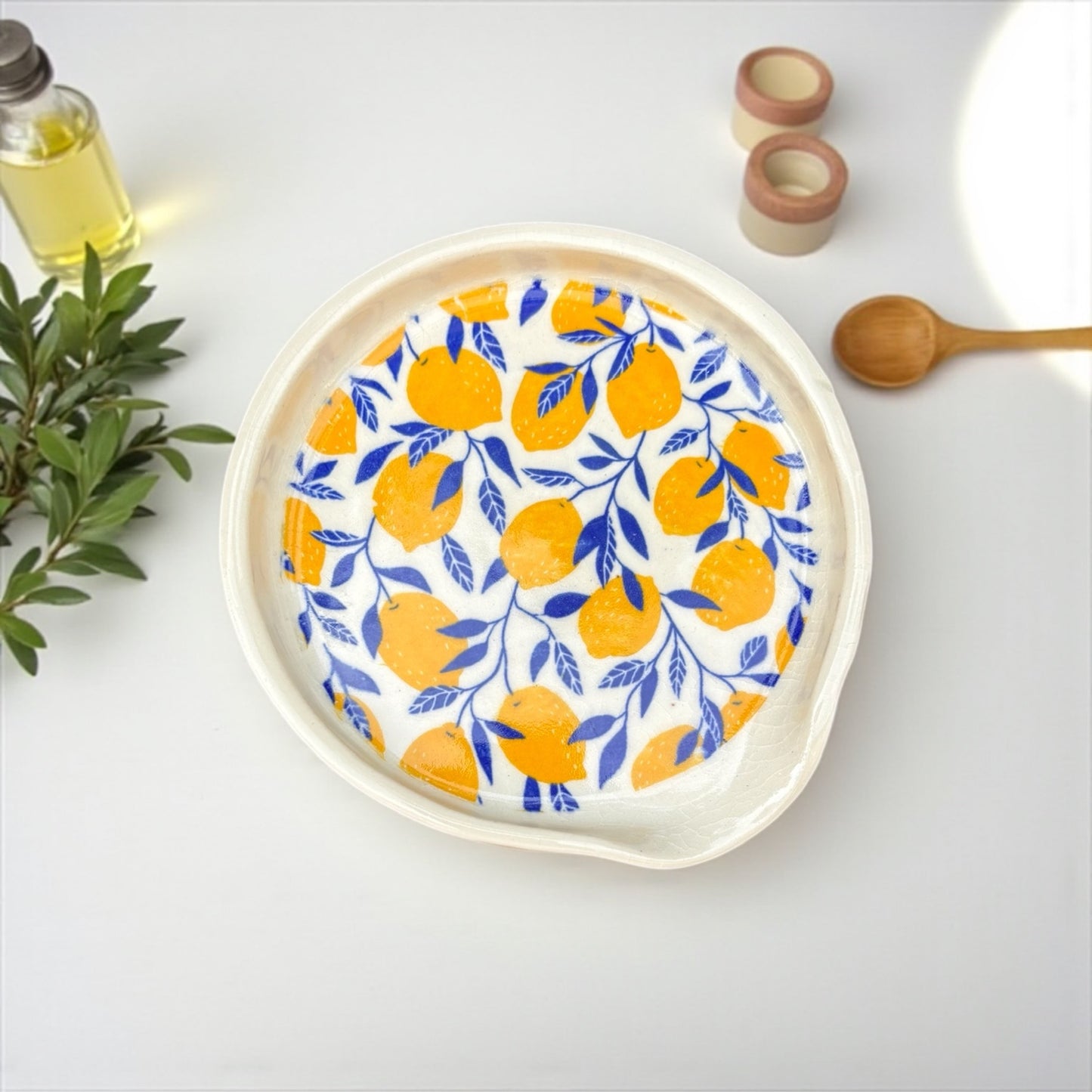 Decorative spoon  rest with lemon pattern on a white surface with a bottle and wooden spoon.